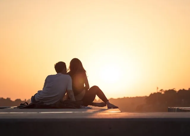 A couple sitting on a pier facing an orange sunset