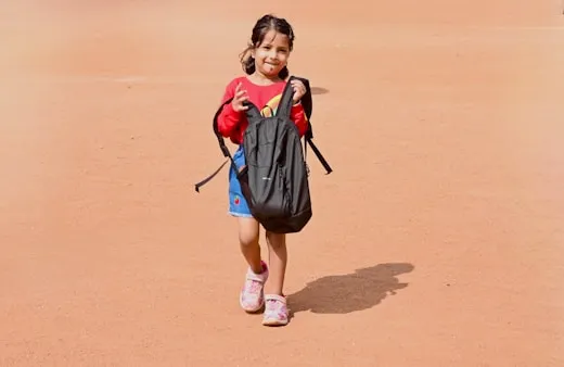 A girl holding a backpack twice her size, smiling